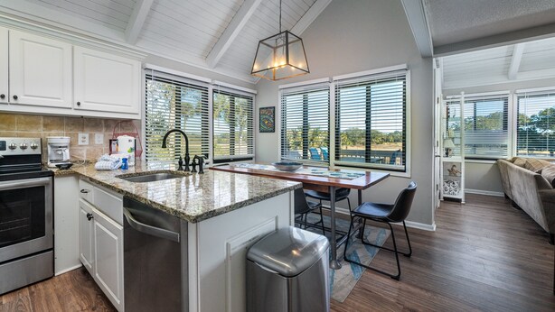 A modern kitchen with white cabinets, a granite countertop, and stainless steel appliances. There is a dining area with a wooden table and chairs, and a large window providing a view of the outdoors. - 2BR/2BA + Loft Cottage! Close to Beach! Golf Course Views! Amenity access Available! (Seabrook Island)