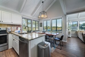 A modern kitchen with white cabinets, a granite countertop, and stainless steel appliances. There is a dining area with a wooden table and chairs, and a large window providing a view of the outdoors. - 2BR/2BA + Loft Cottage! Close to Beach! Golf Course Views! Amenity access Available! (Seabrook Island)