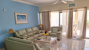 Una sala de estar con un sofá color beige, un sillón a juego, una mesa de centro de vidrio con un jarrón de flores amarillas y un cuadro en la pared.