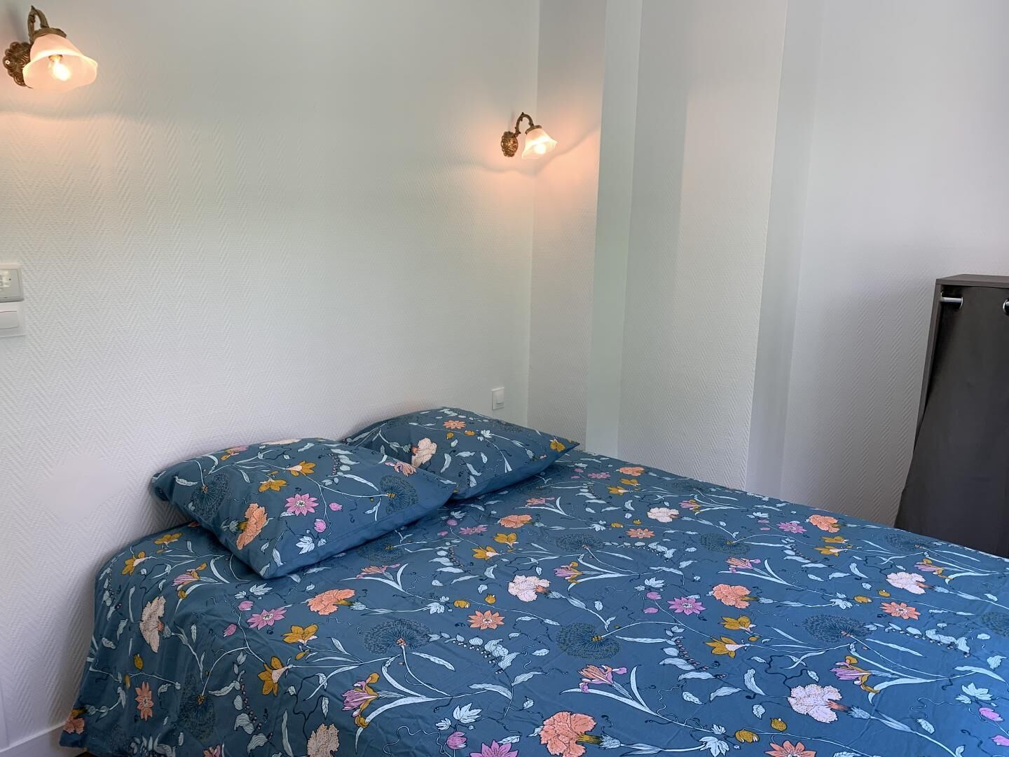 Une chambre à coucher avec une couvre-lit à motifs floraux, une table de chevet et deux appliques murales.