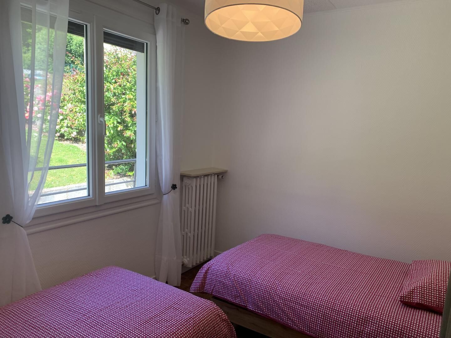 Una habitación con dos camas, una ventana con cortinas translúcidas, un radiador y una lámpara colgante.