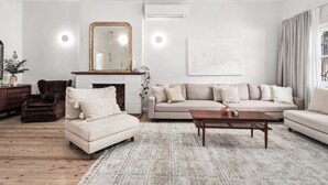 A living room with a beige sofa, a white armchair, a wooden coffee table, and a fireplace with a mirror above it. - Tranquil Coogee Retreat with large backyard & pool (Coogee)