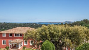 Uma villa vermelha com telhado em telhas, varanda coberta e vista para o mar e colinas.