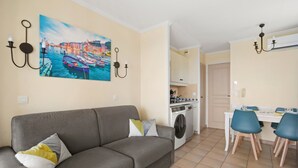 Uma sala de estar com sofá, uma área de refeições com cadeiras, uma máquina de lavar e um quadro na parede.