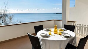 Une table ronde entourée de chaises, sur un balcon donnant sur la mer.