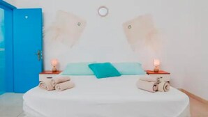Una cama con colchón blanco, dos almohadas y una manta color turquesa. Hay dos mesitas de noche con lámparas, y se ve una puerta azul a la izquierda.