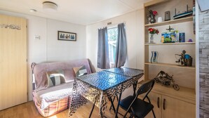 A cozy room with a sofa, a table with a leopard print tablecloth, a chair, a shelf with various items, and a window with curtains. - Mobile home in La Tremblade – Private garden, terrace, playground, and shared pool (La Tremblade (17390))