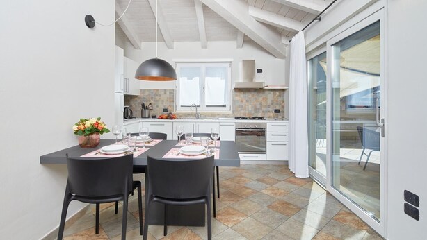 Una cocina moderna con armarios blancos, una zona de comedor con sillas negras y un amplio ventanal corredero al exterior.