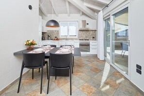 Une cuisine moderne avec des meubles blancs, un coin repas avec des chaises noires et une grande porte-fenêtre coulissante donnant sur l’extérieur.