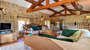 Una habitación rústica con paredes de piedra vista, vigas de madera, una cama con almohadas, un televisor y una mesita de noche con una lámpara.