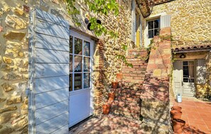 A stone building with a wooden door, a window with lace curtains, and a brick wall with potted plants.
