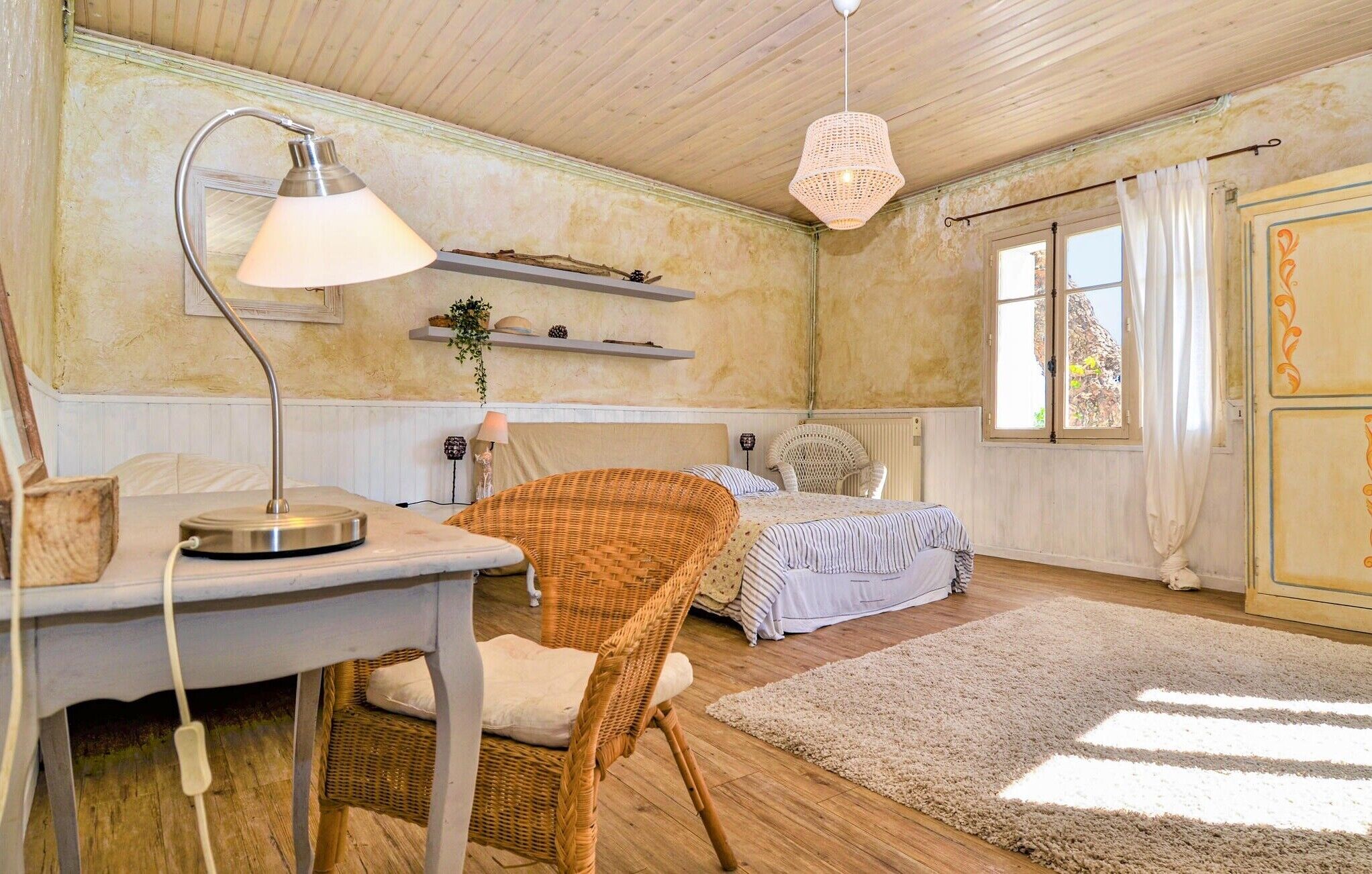 A cozy room with a bed, a wicker chair, a desk with a lamp, and a window with curtains.