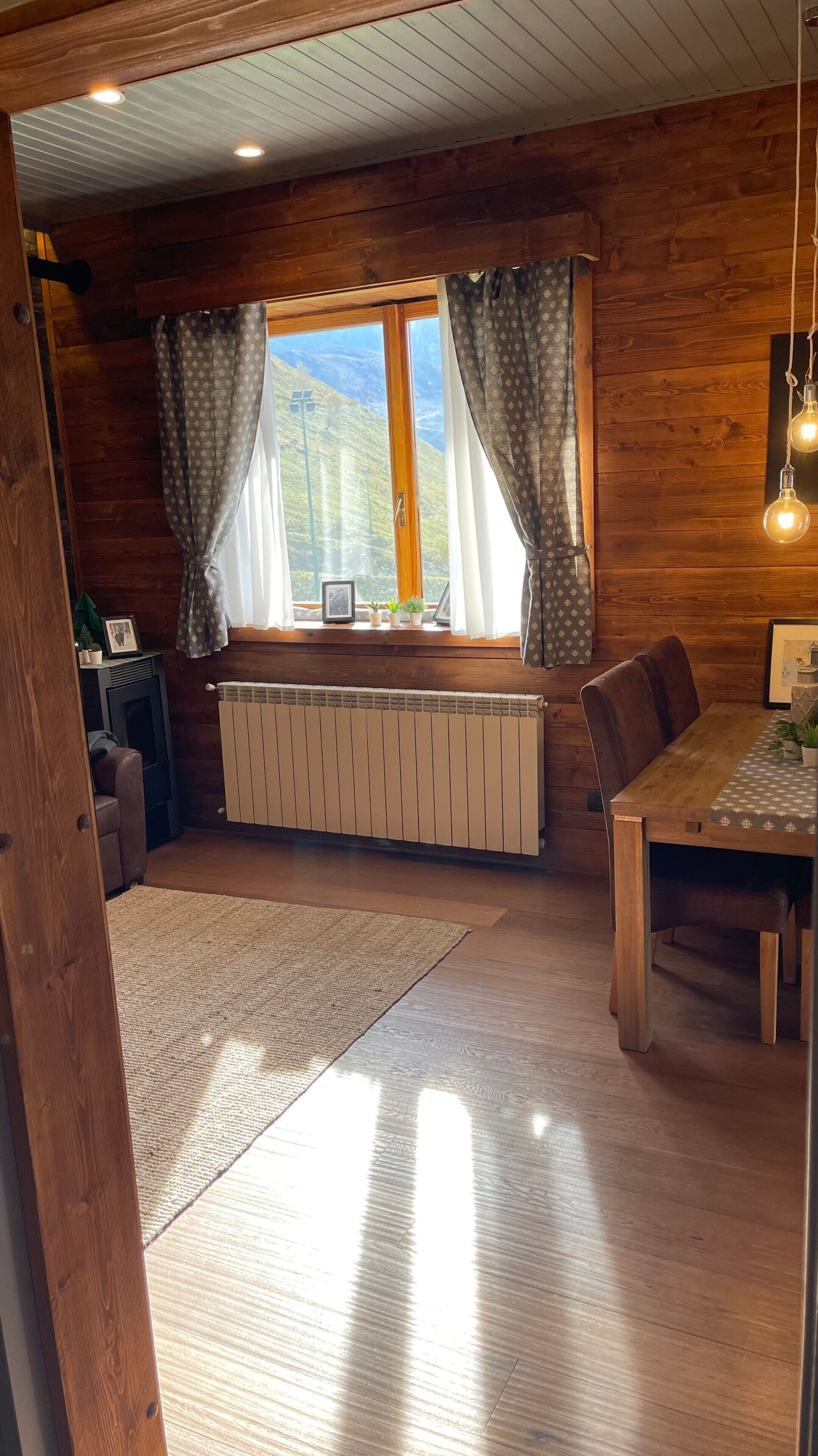 A wooden interior with a dining table, chairs, a radiator, and a window with curtains.