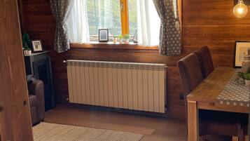 A wooden interior with a dining table, chairs, a radiator, and a window with curtains.