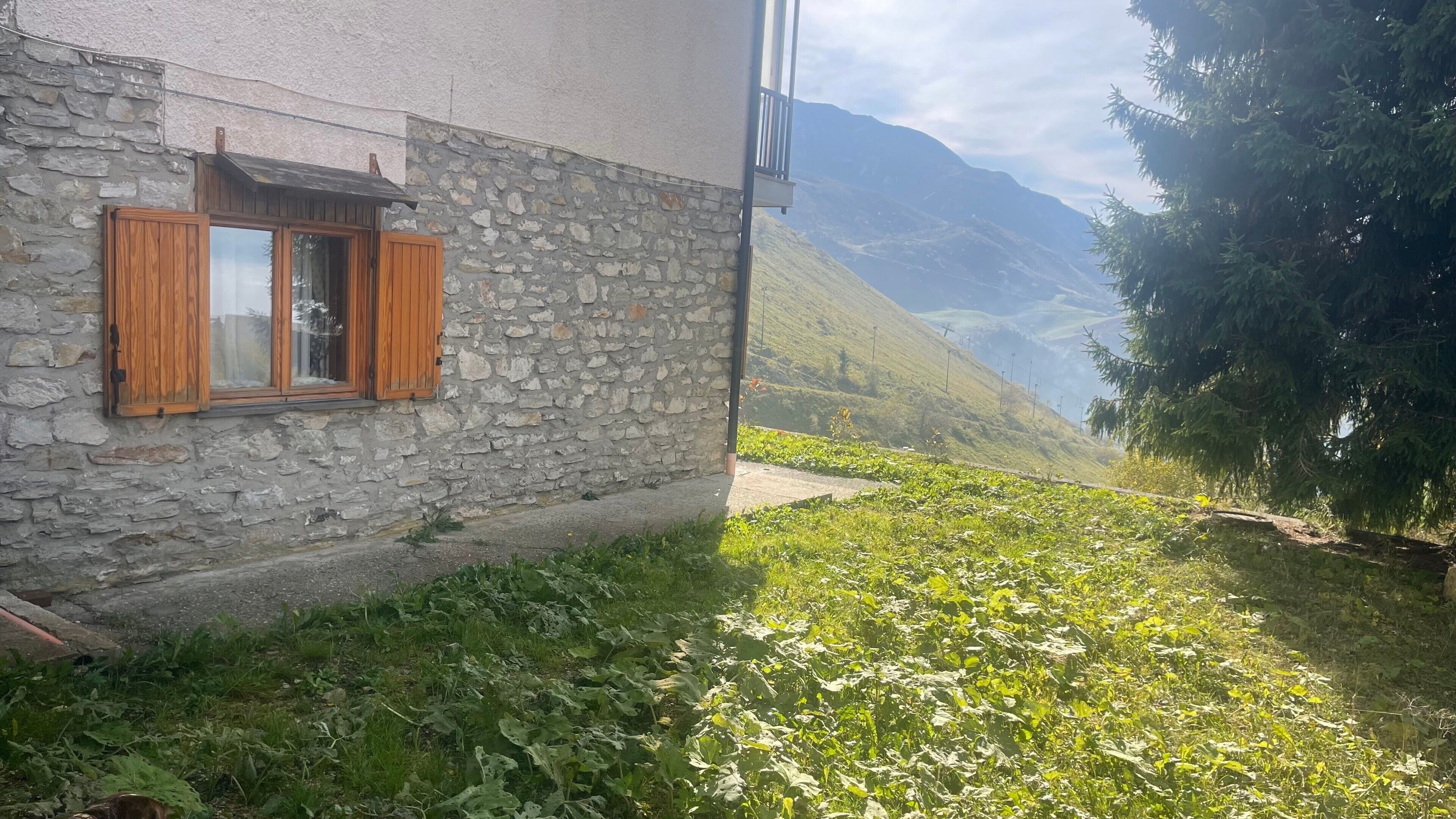 A stone building with wooden shutters, a narrow pathway, and a mountainous backdrop.