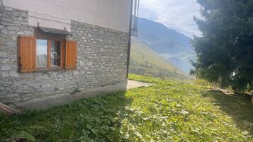 A stone building with wooden shutters, a narrow pathway, and a mountainous backdrop.