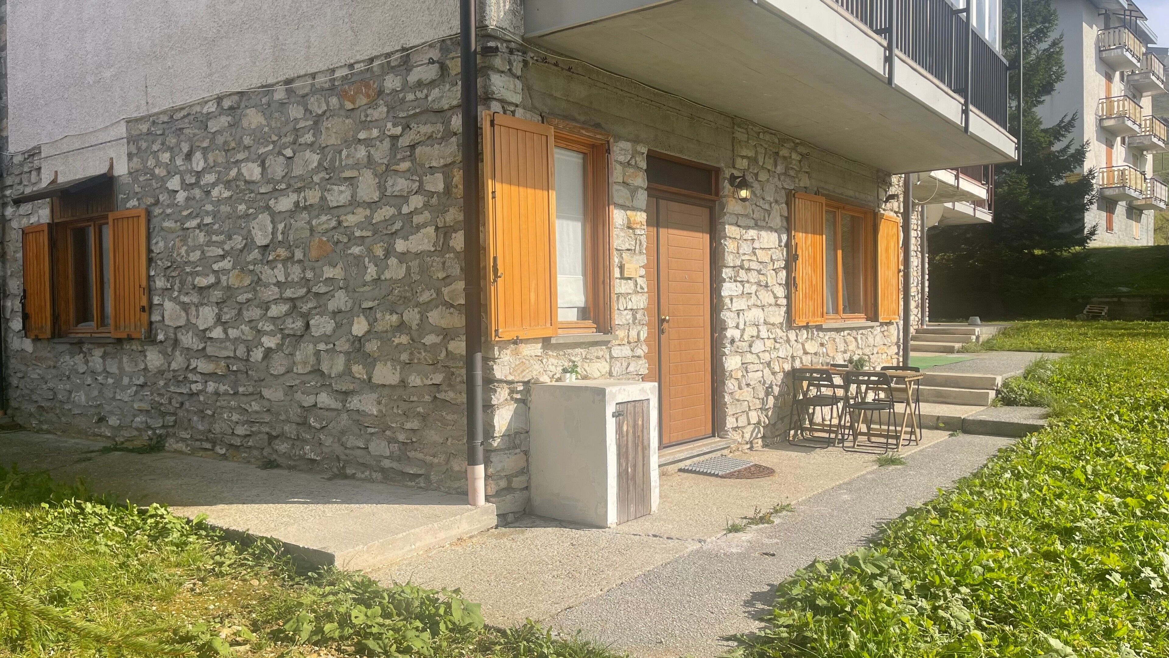 Ein Steinhaus mit hölzernen Fensterläden, einer kleinen Außenbestuhlung und einem Betonweg zum Eingang.