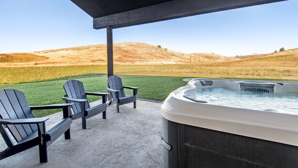 A hot tub and Adirondack chairs on a patio with a view of a grassy field and hills. - Elk Run Ranch With Hot Tub (Tetonia)