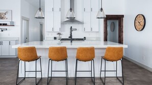 A modern kitchen with white cabinets, a large island with bar stools, pendant lights, and a clock on the wall. - Elk Run Ranch With Hot Tub (Tetonia)