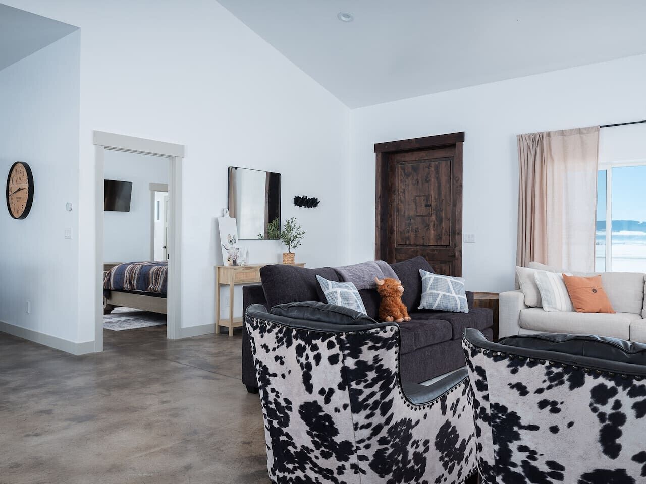 A modern living room with a sofa, a coffee table, and a couch. There is a bed in the background, a mirror on the wall, and a window with curtains.