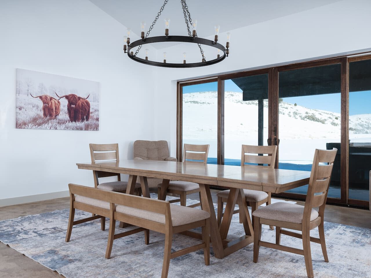 Uma sala de jantar com uma mesa e cadeiras de madeira, um candelabro e uma janela grande com vista para uma paisagem nevada.