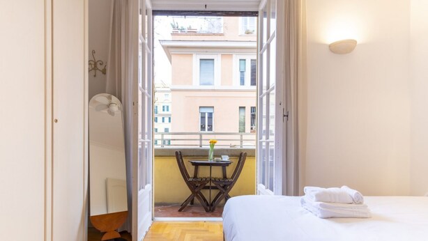 A hotel room with a bed, a small table with a vase, a chair, and a balcony with a view of buildings. - Elegantis Solinda Suite Apartment with Private Terrace - Metro, Tram & Bus (Roma)
