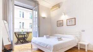 A bedroom with a bed, a small bedside table, a chair, a table with a vase, and a balcony with a view of buildings. - Elegantis Solinda Suite Apartment with Private Terrace - Metro, Tram & Bus (Roma)