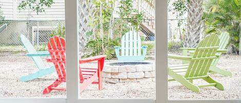 A view through a window of three Adirondack chairs in red, blue, and green, positioned on a gravel area with a stone fire pit in the center.
