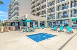 A hotel with a swimming pool, lounge chairs, and a covered area with tables and umbrellas.