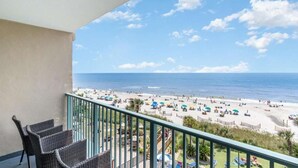 A balcony with wicker furniture overlooking a beach with umbrellas and people.