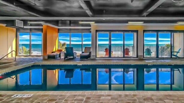 An indoor swimming pool with a view of the ocean, surrounded by lounge chairs and a glass door leading outside.