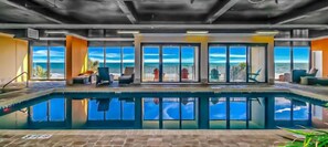 An indoor swimming pool with a view of the ocean, surrounded by lounge chairs and a glass door leading outside.