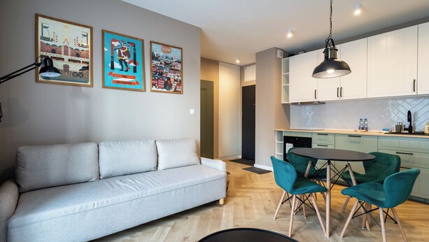 Een moderne woonkamer met een grijze bank, blauwgroene fauteuils en een ronde tafel. In de achtergrond is een keuken met witte kastjes en een hangende lamp zichtbaar.