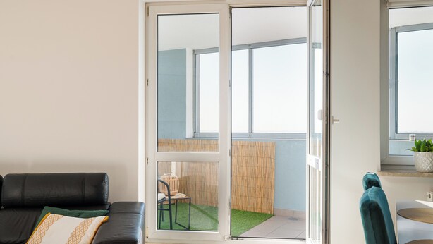 Una sala de estar con un sofá de cuero negro, un cojín verde y un ventanal con vista a un balcón.