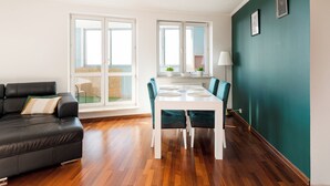 Una sala de estar moderna con un sofá de cuero negro, una mesa de comedor blanca con sillas color turquesa y ventanas grandes con vista a un jardín.