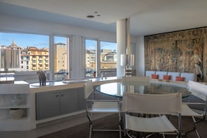 A modern kitchen with a glass island, white chairs, and a view of buildings through large windows.