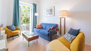 Una sala moderna con un sofá azul, un sillón amarillo y un sofá amarillo con cojines estampados. Hay una mesa de centro, una lámpara y un cuadro en la pared.