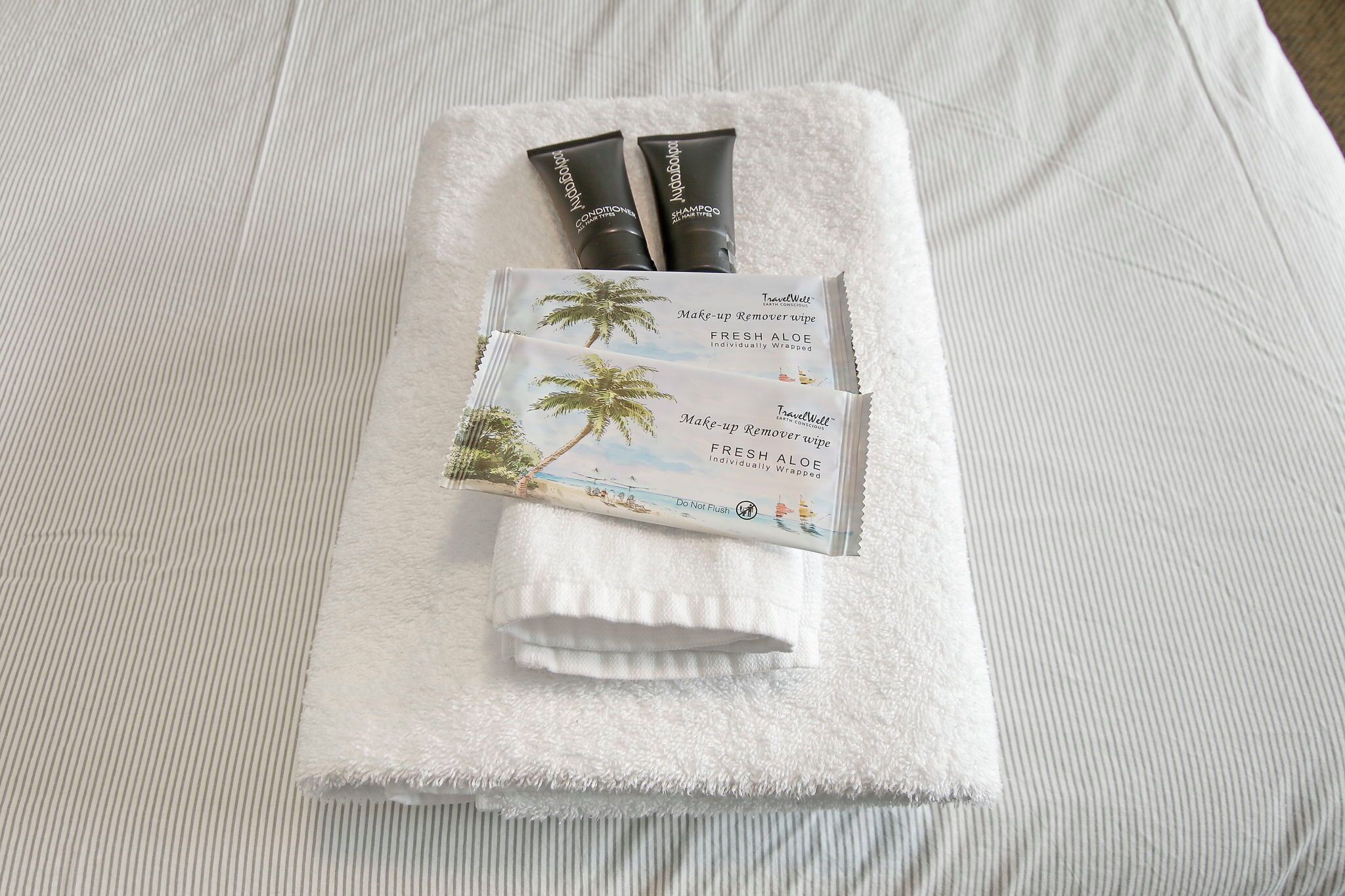 Siististi taitettu valkoinen pyyhe, jonka päällä on kaksi shampootuubia ja tuore aloe-meikkienpoistusliina.