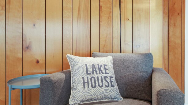 Ein Zimmer mit holzverkleideten Wänden, einem grauen Sofa und einem kleinen blauen Tisch. Auf dem Sofa liegt ein Kissen mit der Aufschrift 'LAKE HOUSE'.
