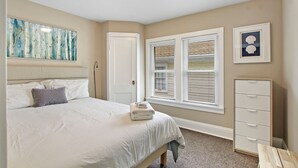 Ein Schlafzimmer mit Bett, Kommode, einem Fenster mit Jalousien und Bildern an den Wänden.