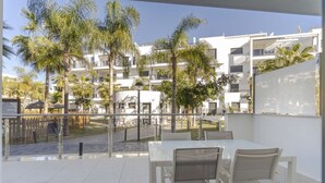 Un balcon avec une table et des chaises blanches, donnant sur une cour intérieure agrémentée de palmiers et sur un immeuble doté de balcons.