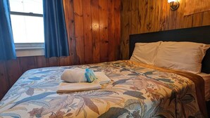A room with wooden panel walls, a bed with floral bedding, a nightstand, and a window with curtains.