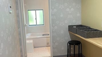 A small, compact room with a wooden floor, a desk with a stool, a window, and a door leading to another room.