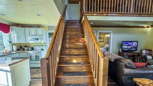 A wooden staircase leading to a second floor, a kitchen with white cabinets, a dining area with a leather couch, and a television in the living room. - New: Charming and Cozy, Great Value! (Swanton)