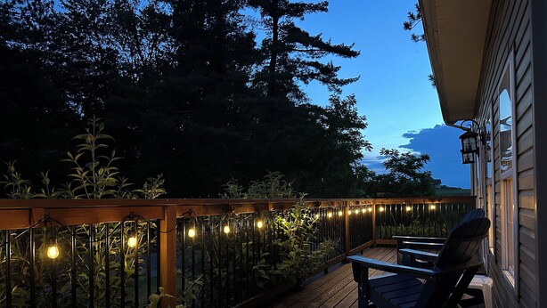 A wooden deck with string lights, a rocking chair, and a view of trees and a house. - New: Charming and Cozy, Great Value! (Swanton)