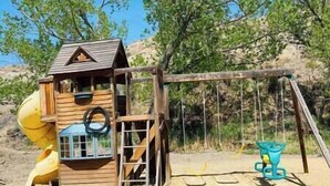 Una estructura de juego de madera con un tobogán y columpios, ubicada en un entorno natural al aire libre.