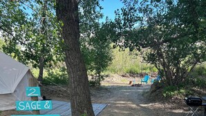 Un camping con una tienda de campaña, un letrero que dice 'SAGE & WILDFLOWER' y un camino de grava que lleva a una zona de estar con sillas.
