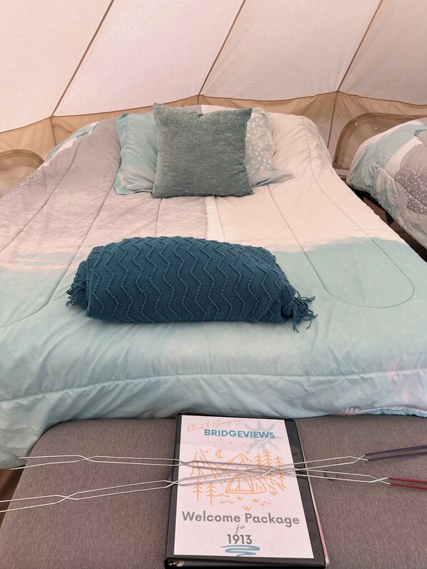 Una cama con una manta y almohadas azules, un paquete de bienvenida y una estructura parecida a una tienda de campaña al fondo.