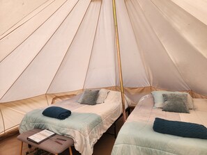 A tent with two beds, each with a pillow and a blanket. There is a small table with a book and a pair of glasses.