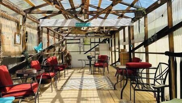 A covered outdoor seating area with red chairs and tables, a patterned rug, and a wooden ceiling structure.
